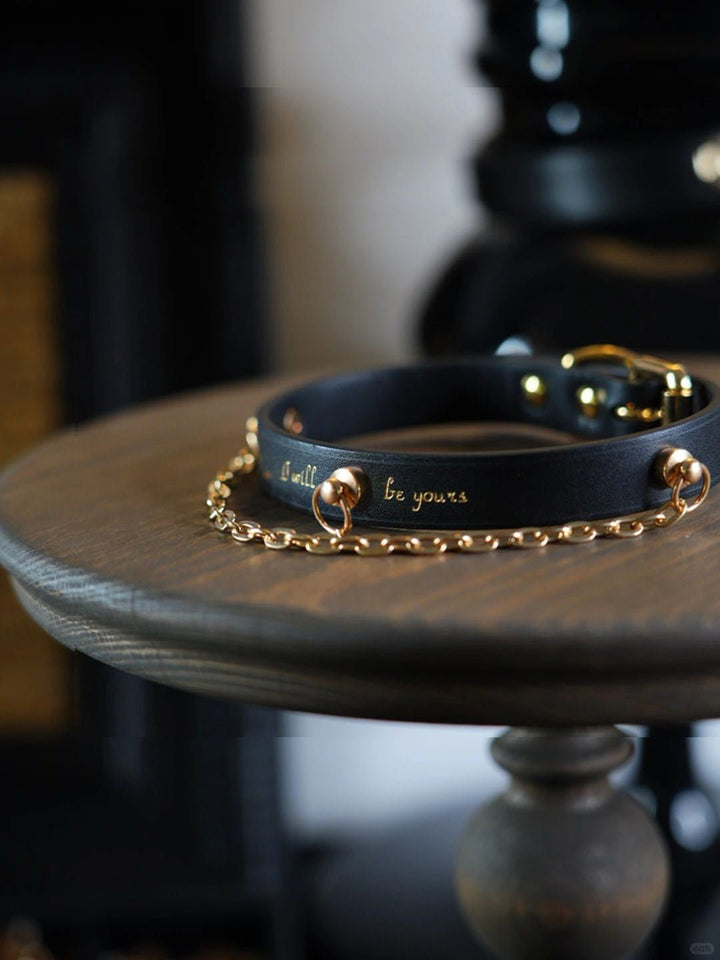 A black leather collar with gold chain accents and engraved with the words "I will be yours." This elegant BDSM-inspired collar features a sleek design with gold hardware, including D-rings for leash attachments.