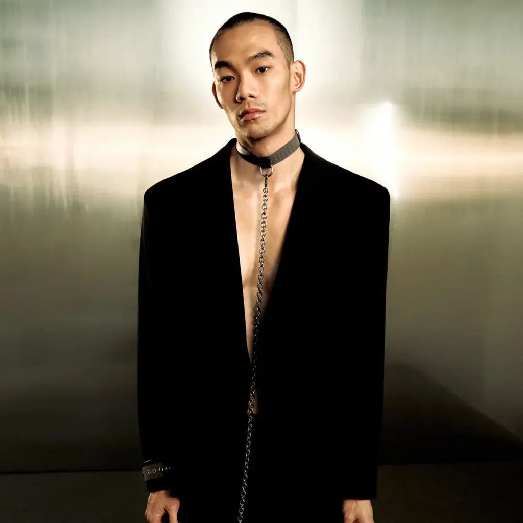 A man wearing a black tailored suit with a very low open chest, a black choker necklace with a hanging chain, and a black bracelet, standing against a dark metallic studio background. The text 'BOUND | 无隙' appears at the bottom.