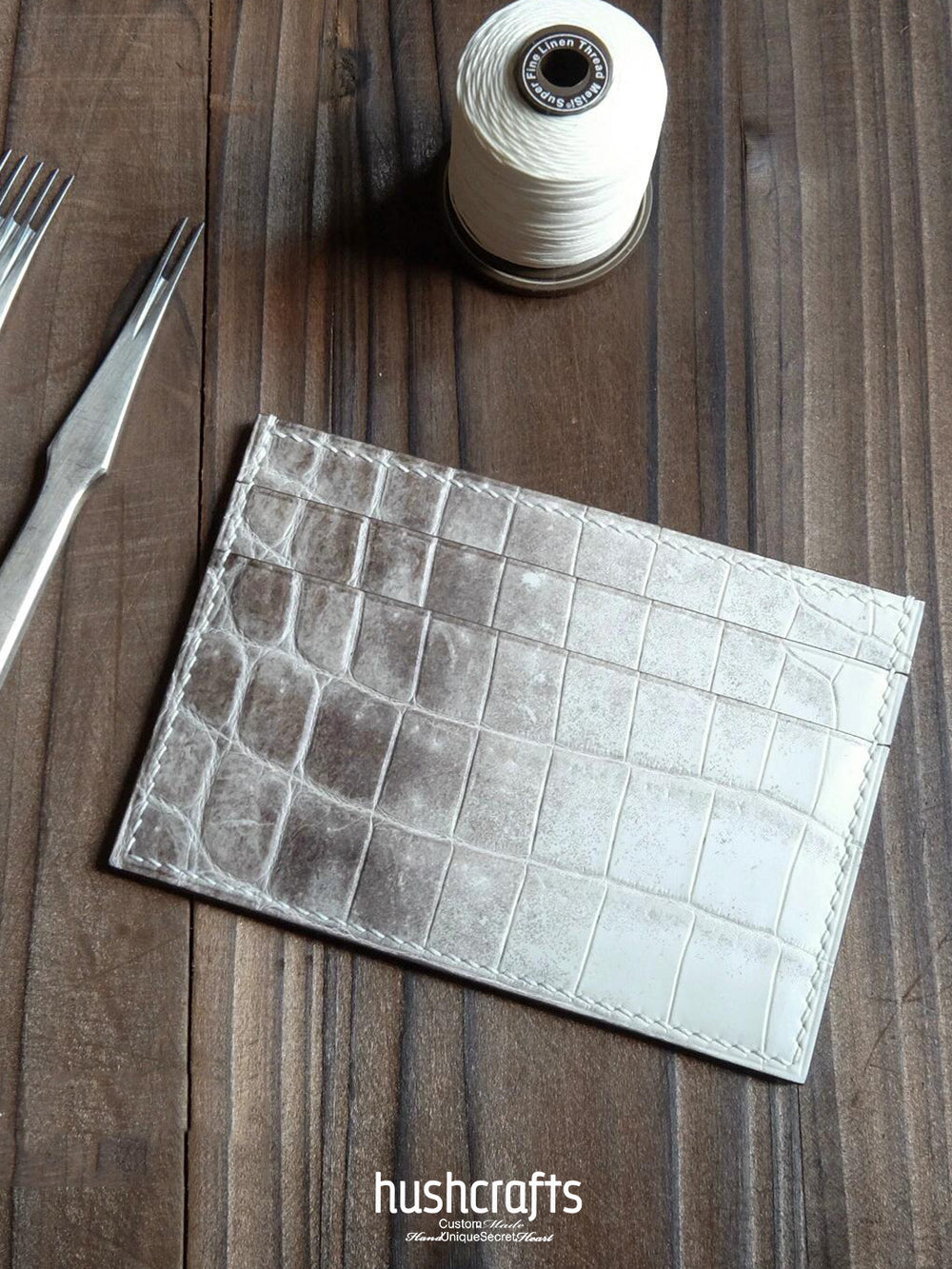 Crocodile texture leather card on a wooden surface with thread and fork, featuring 'hushcrafts' branding.