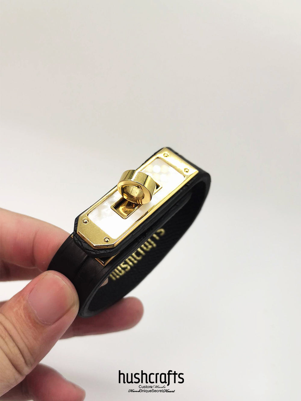 Black leather bracelet with gold clasp held by a hand on a white background, featuring the brand 'hushcrafts'.