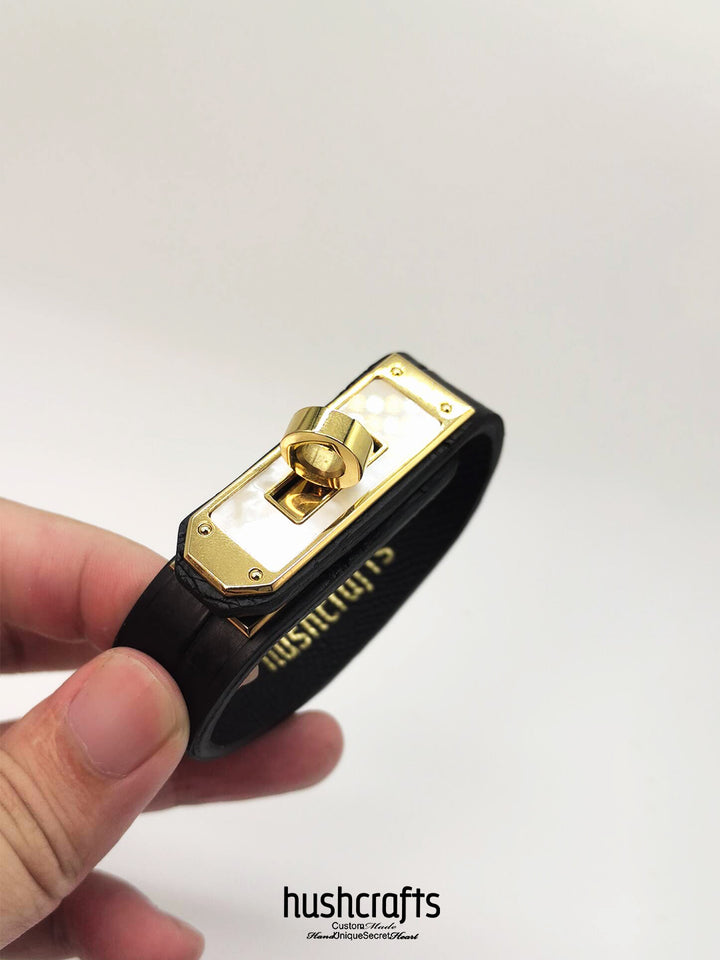 Black leather bracelet with gold clasp held by a hand on a white background, featuring the brand 'hushcrafts'.