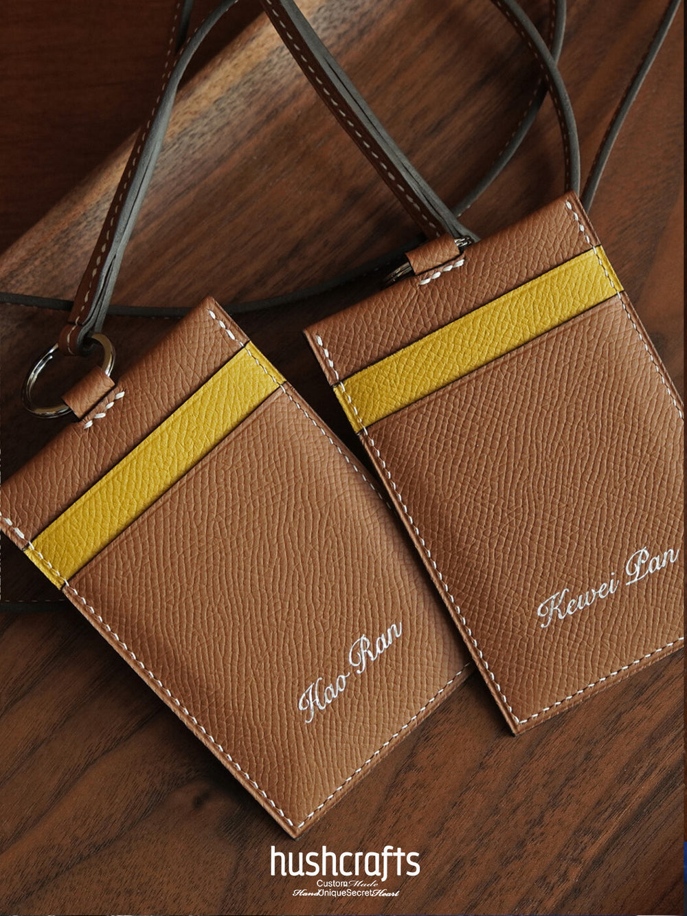 Two brown leather card holders with yellow stripes on a wooden surface, featuring the brand 'hushcrafts'.