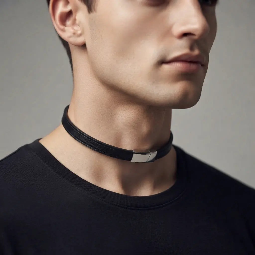 A close-up of a man wearing a black leather choker collar with a polished silver metal accent clasp, paired with a black crew-neck t-shirt against a neutral gray background.