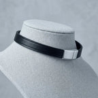 A black leather choker collar with a polished silver metal rectangular clasp, displayed on a light gray fabric jewelry bust against a textured gray background.