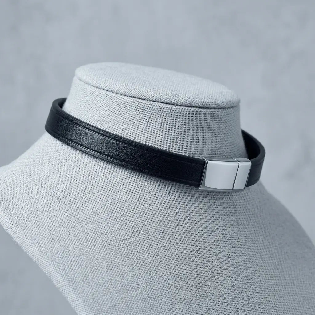 A black leather choker collar with a polished silver metal rectangular clasp, displayed on a light gray fabric jewelry bust against a textured gray background.