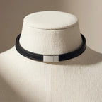 A black leather choker collar with a polished silver rectangular clasp, displayed on a cream-colored fabric jewelry mannequin bust.