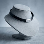 A black leather band with a silver metal clasp, displayed on a gray fabric jewelry bust against a gray background.