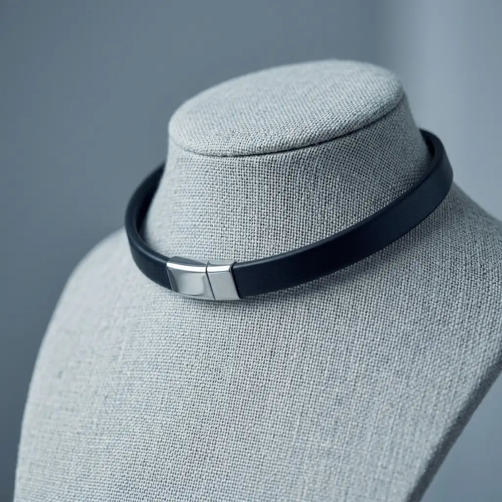 A black leather choker collar with a silver-toned rectangular metal clasp, displayed on a gray fabric jewelry bust against a soft gray background.