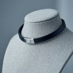 A black leather choker collar with a silver-toned rectangular metal clasp, displayed on a gray fabric jewelry bust against a soft gray background.