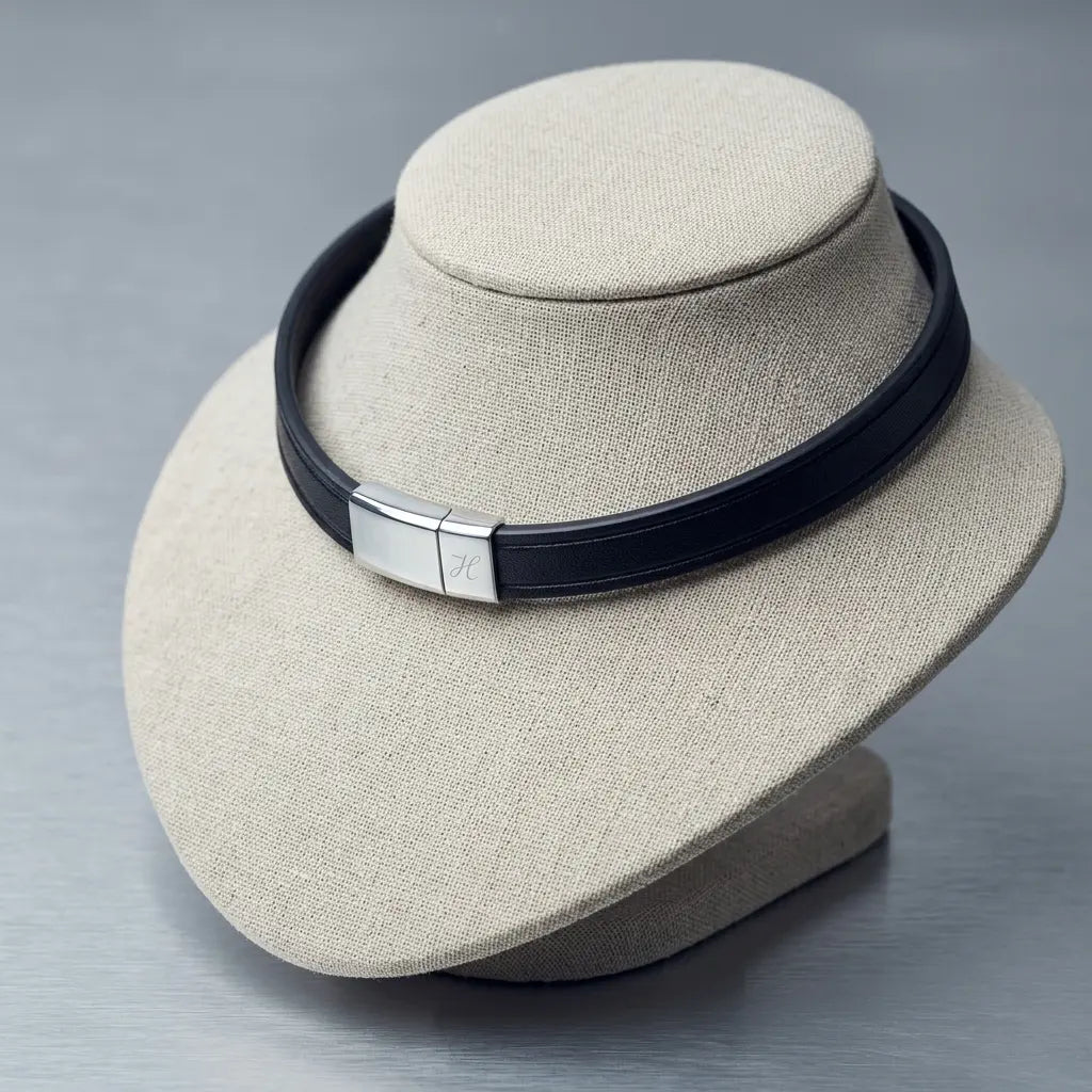 A black leather collar with a polished silver-toned metal clasp, displayed on a beige fabric jewelry bust against a gray background.