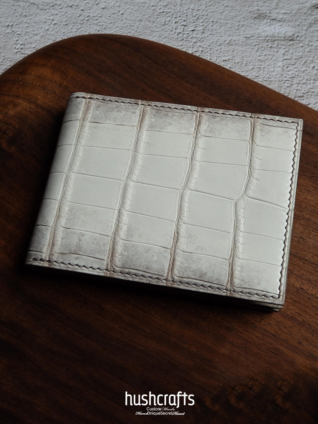 White leather wallet with crocodile texture on a wooden surface, featuring the brand 'hushcrafts'.