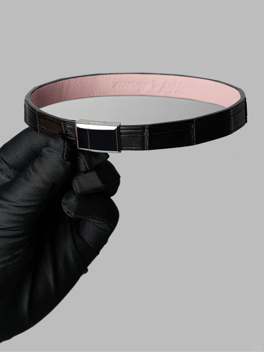 Black crocodile leather choker with pink lambskin lining and a stainless steel magnetic clasp, held in a black-gloved hand.