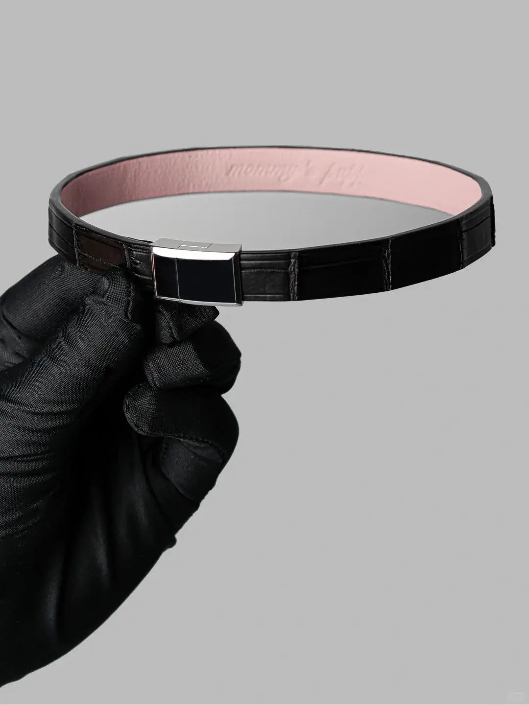 Black crocodile leather choker with pink lambskin lining and a stainless steel magnetic clasp, held in a black-gloved hand.